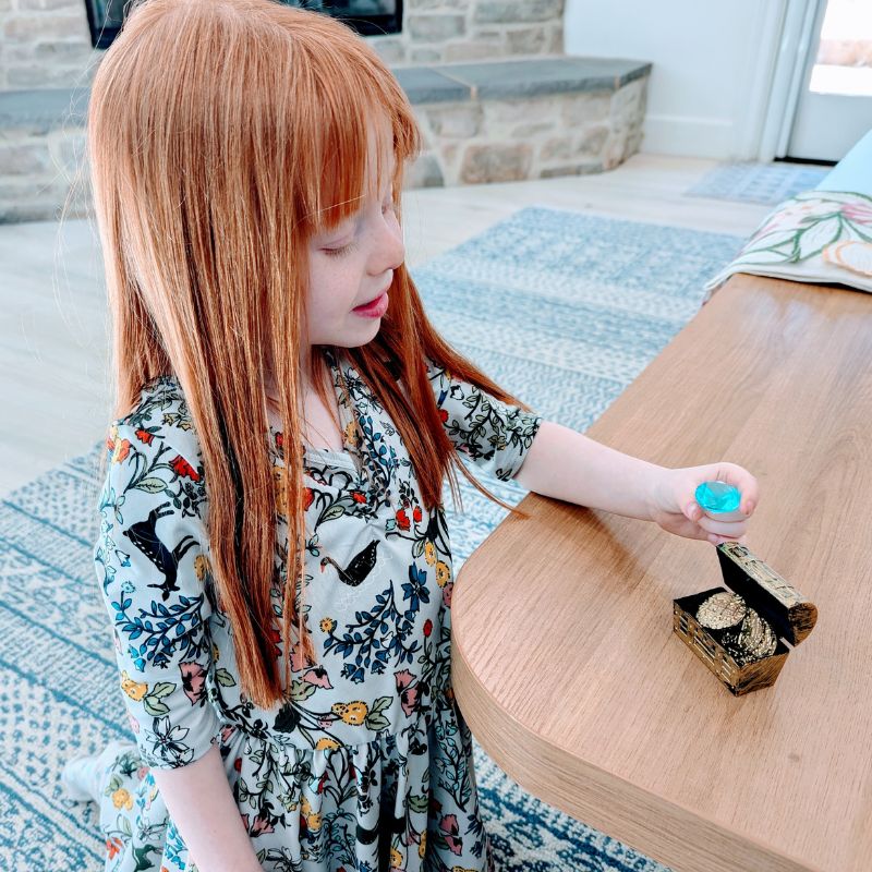 Girl holding a blue gem next to a treasure chest with gold coins.