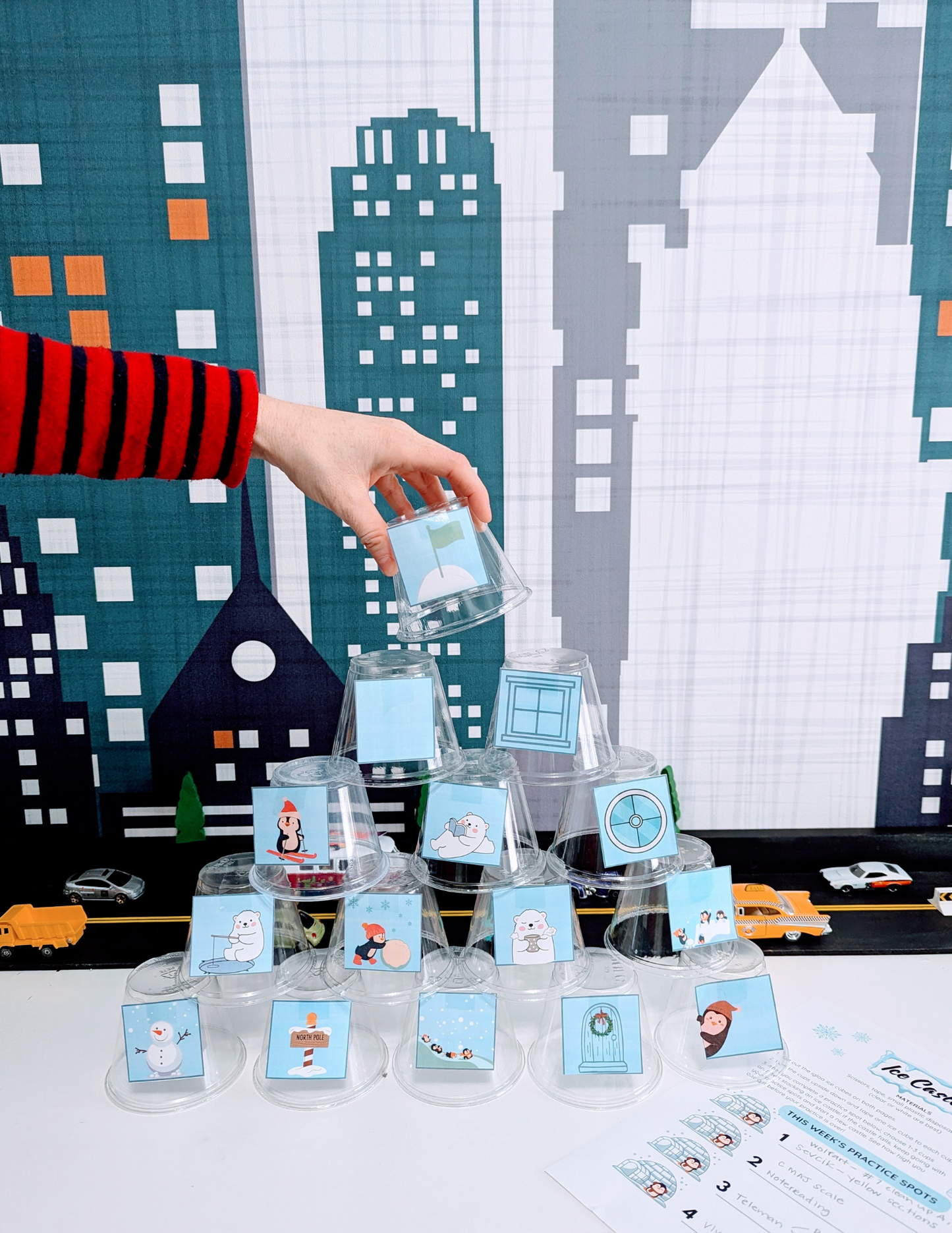 A person's hand places a clear cup on the top of a cup tower that has winter images